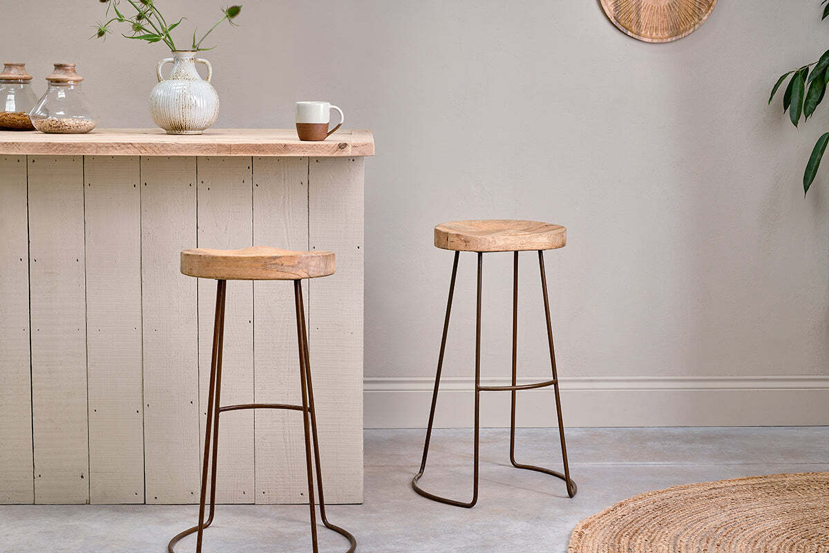 Two Loko Mango Wood Stools by nkuku, with dark iron legs and sustainable mango wood seats, stand in front of a light wood bar. A textured white vase with greenery and a two-tone mug complete the inviting scene.
