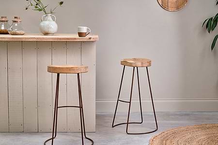 A minimalist kitchen showcases a light wooden counter with jars, a vase, and a mug. Two nkuku Loko Mango Wood Stools - Tall add rustic charm with iron frames and wooden seats, while a woven rug and potted plant accent the neutral decor.