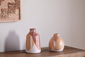 Two nkuku Lucia Ceramic Vases in a terracotta mix with geometric patterns rest on a wooden surface against a light wall. A framed woven textile to the left adds texture to the minimalist decor.