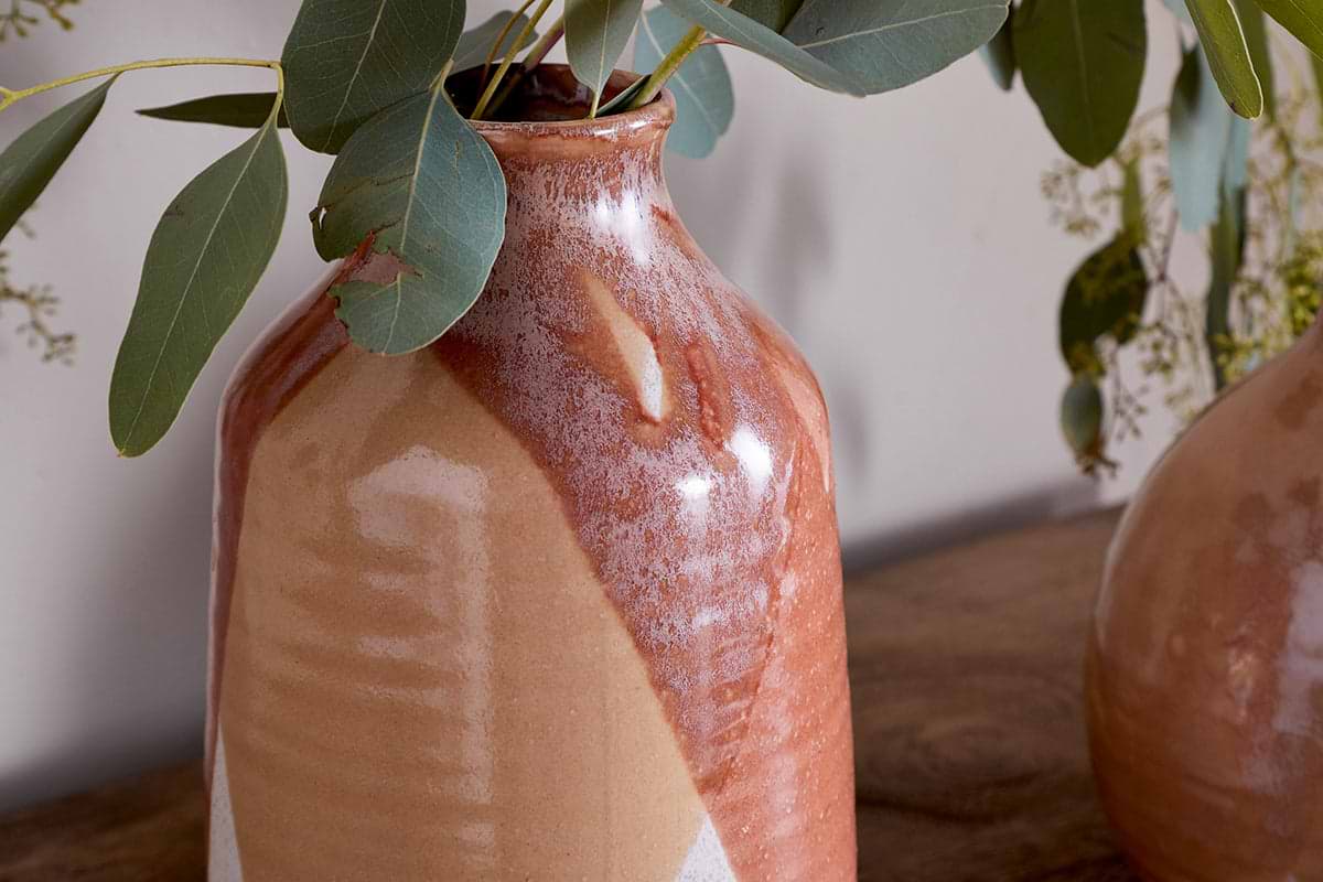 A close-up of the nkuku Lucia Ceramic Vase - Terracotta Mix, featuring a glossy marbled glaze. Green eucalyptus leaves extend from the vase, with another similar vase on a wooden surface and a light-colored wall in the background.