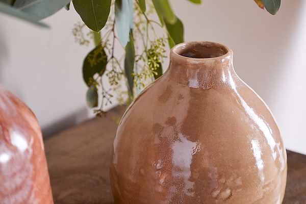A close-up of the nkuku Lucia Ceramic Vase - Terracotta Mix sits on a wooden surface. Green eucalyptus leaves and yellow buds hang above, while a softly blurred background highlights the vase’s glossy finish.