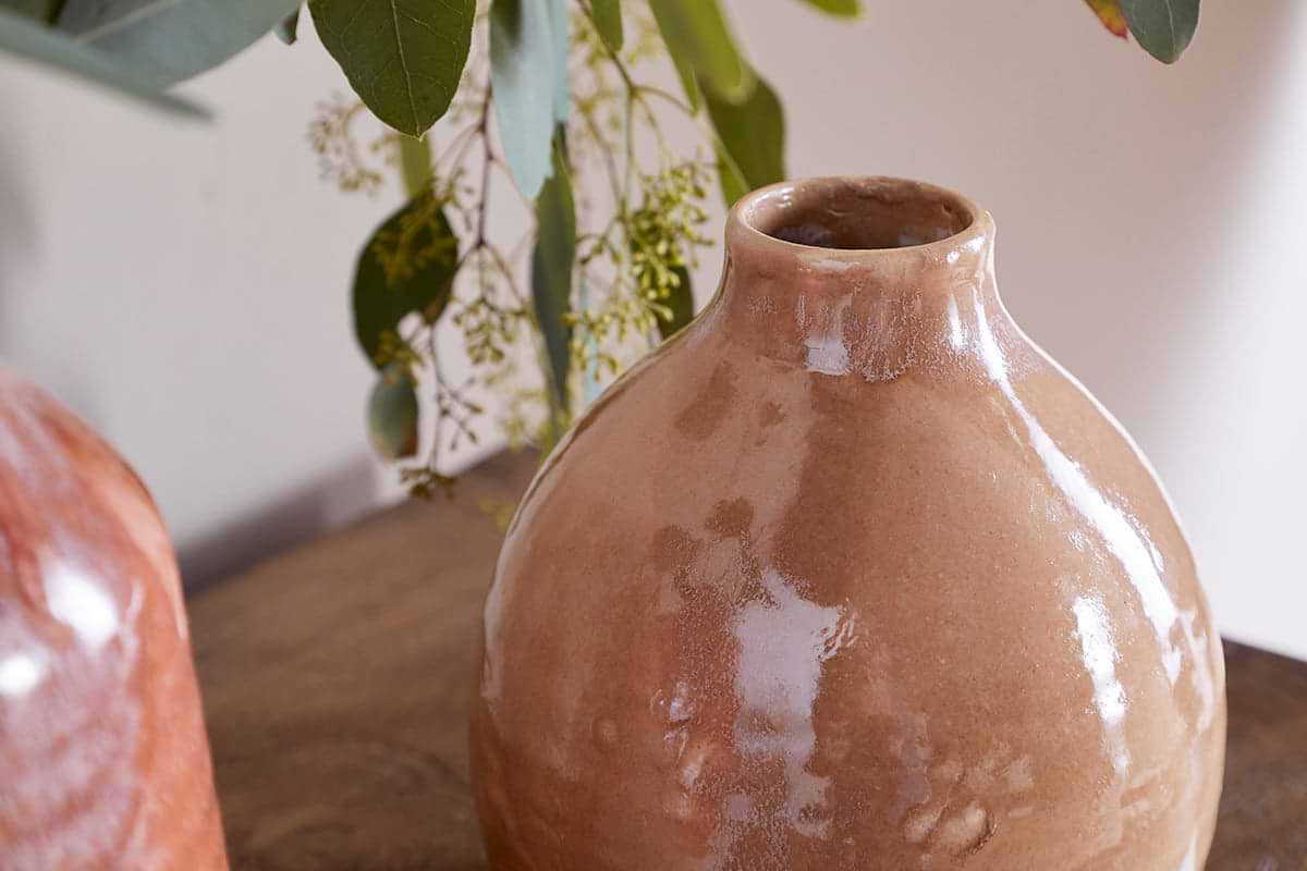A close-up of the nkuku Lucia Ceramic Vase - Terracotta Mix sits on a wooden surface. Green eucalyptus leaves and yellow buds hang above, while a softly blurred background highlights the vase’s glossy finish.
