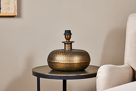 The Lumbu Lamp - Antique Brass - Small by nkuku, with its metallic textured artisan base, sits perfectly on a side table next to a beige chair, adding stylish decorative lighting to any space.