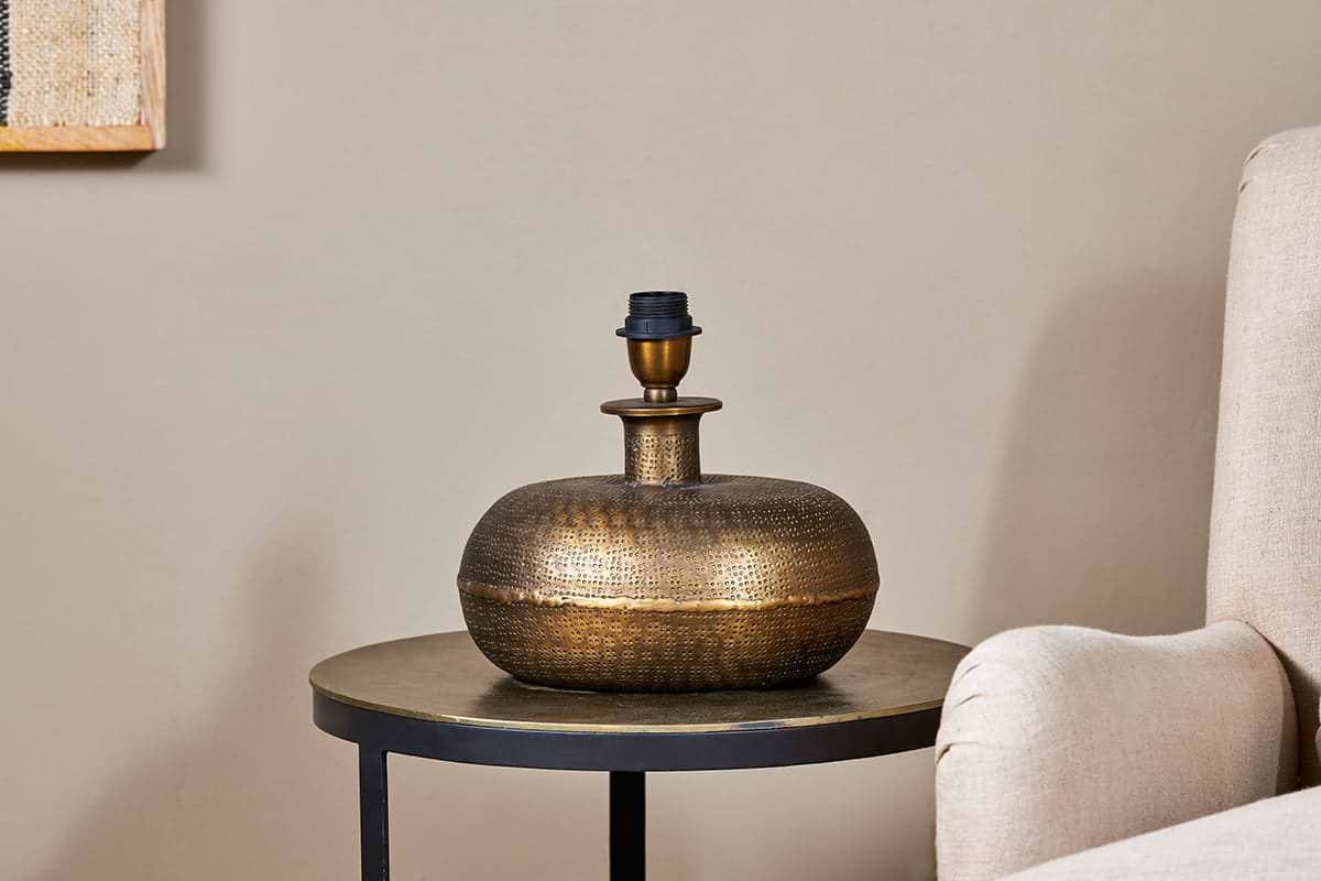 The Lumbu Lamp - Antique Brass - Small by nkuku, with its metallic textured artisan base, sits perfectly on a side table next to a beige chair, adding stylish decorative lighting to any space.