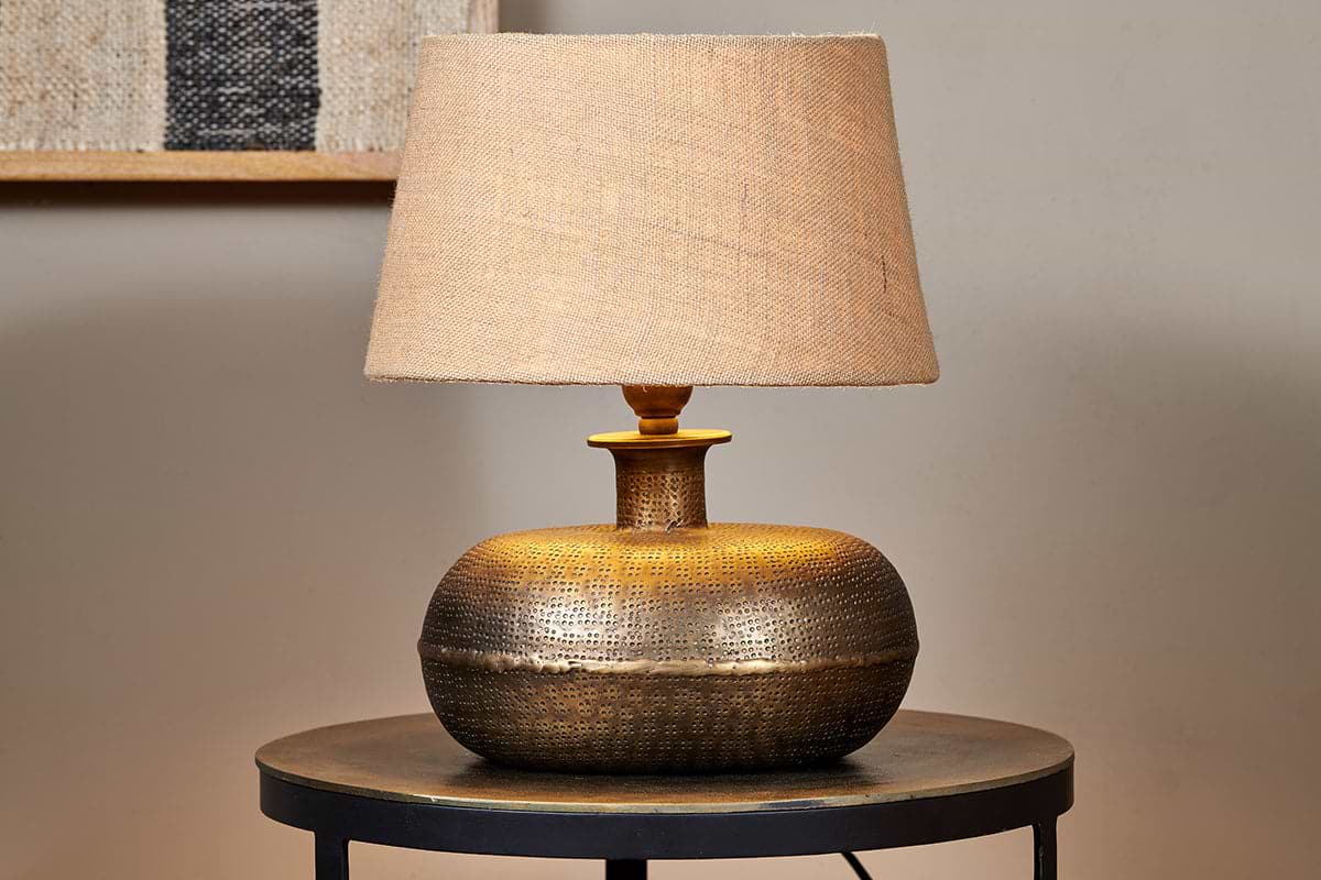 The nkuku Lumbu Lamp in Antique Brass (Small) with a wide, textured base and beige fabric shade sits on a round, dark side table against a neutral wall. Turned on, it casts warm decorative light. A picture frame is visible in the background.