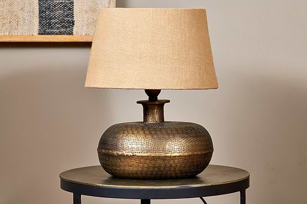 The nkuku Lumbu Lamp in antique brass, small size, with a textured base and beige fabric shade sits on a metallic side table. A neutral wall and framed artwork in the background highlight this elegant decorative lighting piece.