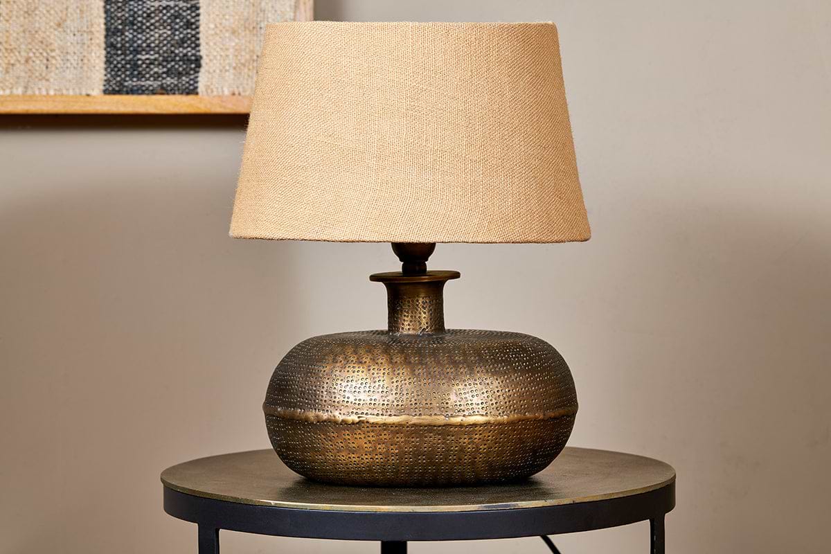 The nkuku Lumbu Lamp in antique brass, small size, with a textured base and beige fabric shade sits on a metallic side table. A neutral wall and framed artwork in the background highlight this elegant decorative lighting piece.