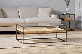 The nkuku Luzon Iron & Mango Wood Display Coffee Table – Brass sits on a woven rug in front of a sectional sofa, with a side table featuring wooden shelves in the background.
