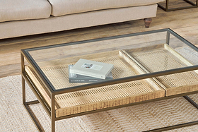 The nkuku Luzon Iron & Mango Wood Display Coffee Table in brass stands on a beige textured rug with stacked books and mango wood trays. A beige sofa with wooden legs sits in the background on a wooden floor.