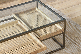 The nkuku Luzon Iron & Mango Wood Display Coffee Table in brass features a clear glass top, black metal frame, and two sliding mango wood trays with woven rattan bottoms, shown partially open on a textured beige rug over a wooden floor.