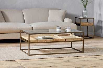 A modern living room featuring the nkuku Luzon Iron & Mango Wood Display Coffee Table in brass, topped with mango wood trays, striped cups, and books. A small side table holds a glass vase and plant over a light rug on wooden floors.