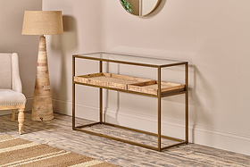 The nkuku Luzon Iron & Mango Wood Display Console Table features a modern brass frame, clear glass top, and two woven baskets on a lower shelf. It's styled against a beige wall with a floor lamp, upholstered chair, and rug on wood-patterned flooring.