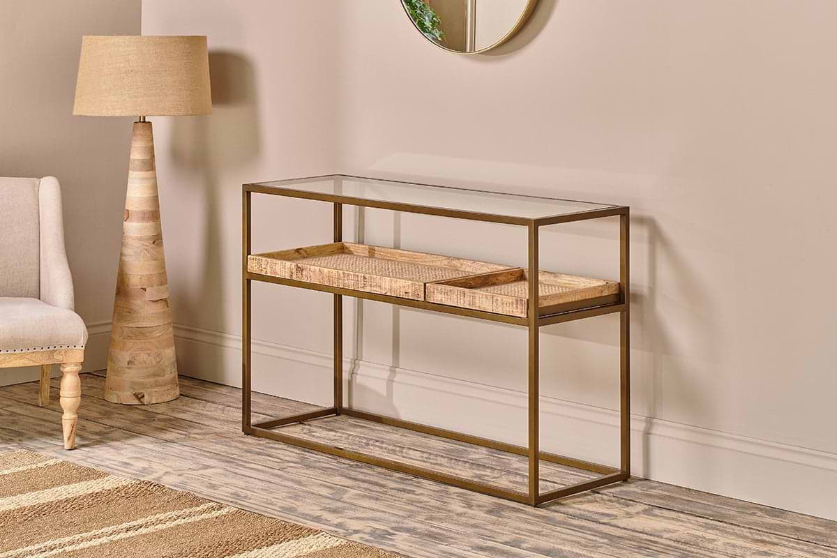 The nkuku Luzon Iron & Mango Wood Display Console Table features a modern brass frame, clear glass top, and two woven baskets on a lower shelf. It's styled against a beige wall with a floor lamp, upholstered chair, and rug on wood-patterned flooring.