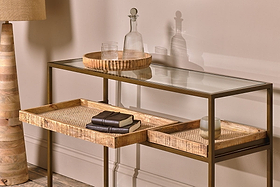 The Luzon Iron & Mango Wood Display Console Table by nkuku holds two woven trays—one with notebooks. Above, a round wooden tray displays a glass decanter and tumblers. A lamp with a wooden base stands nearby against a beige wall.