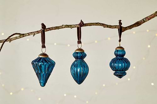Three indigo glass Manipur Baubles by nkuku, featuring ribbed textures and brass tops, hang from dark ribbons. The set of three ornaments—diamond, teardrop, and round—adds vintage charm to your festive decor.