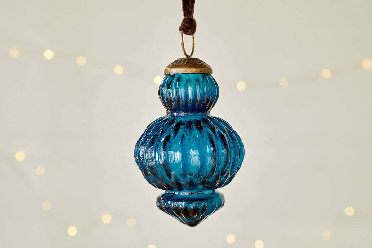 The nkuku Manipur Baubles - Indigo (Set of 3) feature blue ribbed glass with brass caps and loops, hanging from brown cords. Their vintage style and warm glow evoke classic festive charm against a cream background.