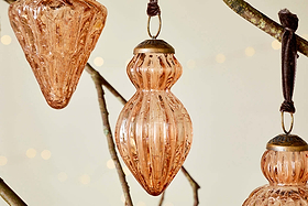 Three ribbed glass Manipur Baubles - Copper (Set of 3) by nkuku, each with bronzed tops and brown ribbons, hang from a branch. Their vintage style and warm tones stand out against softly lit festive decor and twinkling lights.