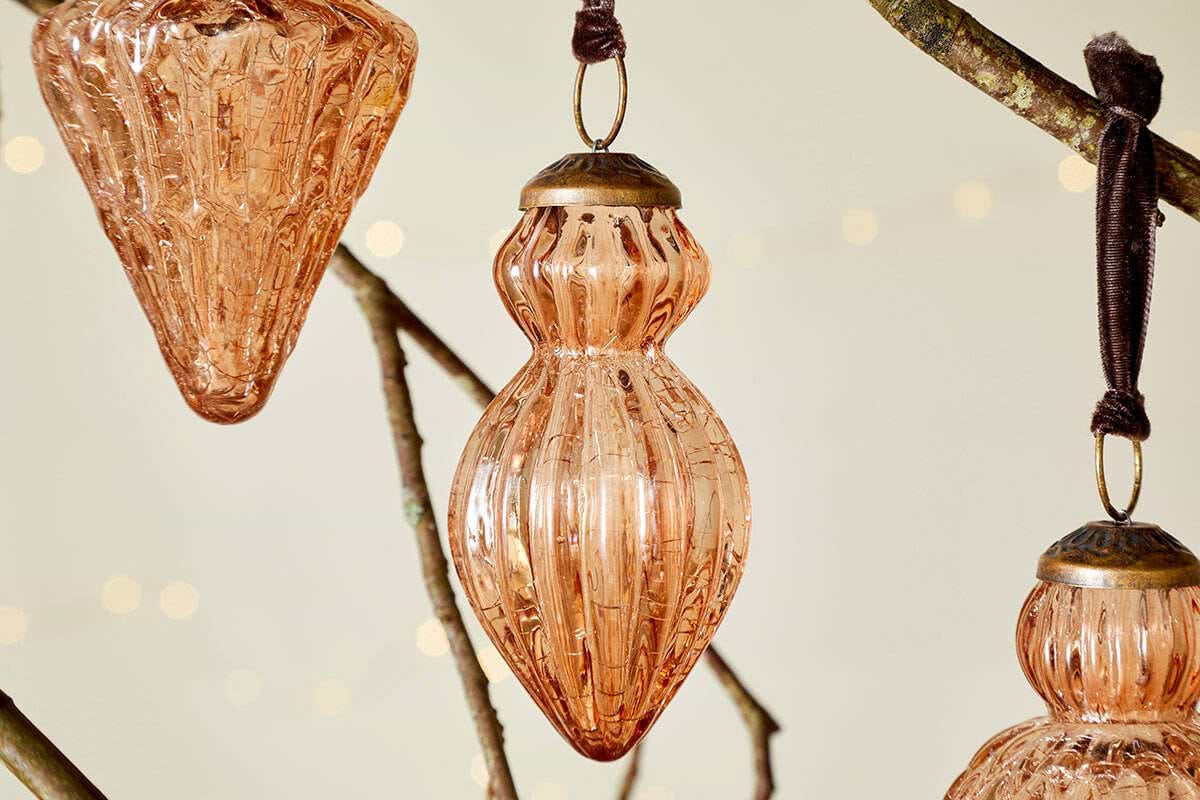 Three ribbed glass Manipur Baubles - Copper (Set of 3) by nkuku, each with bronzed tops and brown ribbons, hang from a branch. Their vintage style and warm tones stand out against softly lit festive decor and twinkling lights.