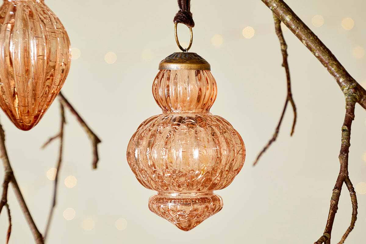 The Manipur Baubles - Copper (Set of 3) by nkuku feature ribbed glass and brass caps. Hanging from brown strings on bare branches, they evoke vintage charm with warm fairy lights glowing softly in the background for a festive touch.