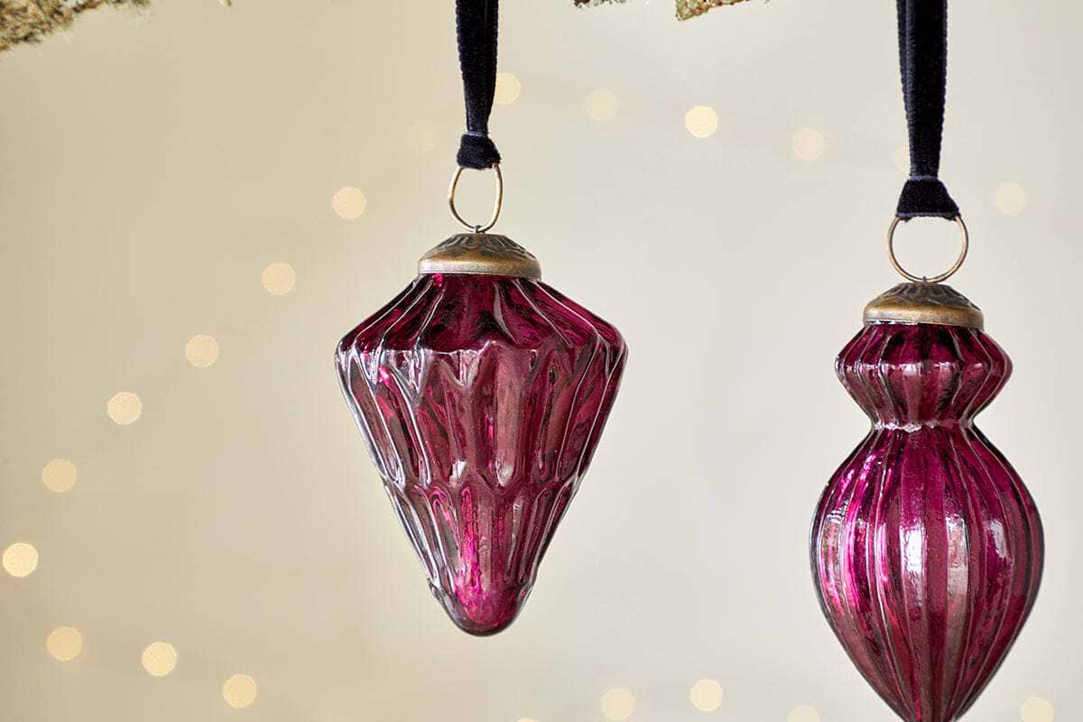Three vintage-style Manipur Baubles - Ruby by nkuku, crafted from deep magenta glass, hang from black velvet ribbons. Each ornament features a gold-colored cap and glows beautifully against softly blurred fairy lights.