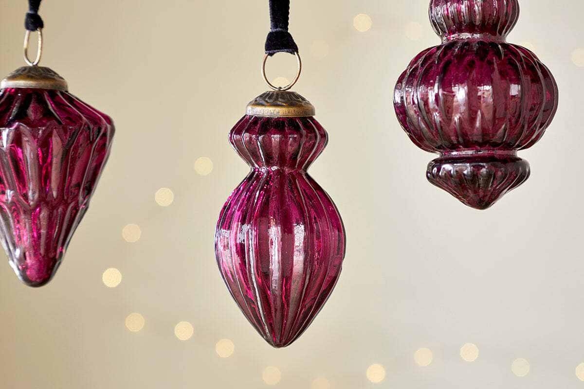 The nkuku Manipur Baubles - Ruby (Set of 3) feature ribbed, vintage-style magenta glass ornaments on black ribbons, displayed against a cream background with golden lights for an elegant festive touch.
