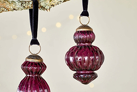 Three vintage-style Manipur Baubles - Ruby by nkuku, featuring rich ruby glass and gold tops, hang from black velvet ribbons on a tree branch, adding classic charm to festive décor against a neutral backdrop with soft warm lights.