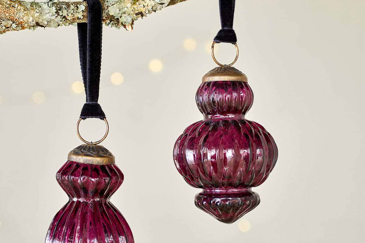 Three vintage-style Manipur Baubles - Ruby by nkuku, featuring rich ruby glass and gold tops, hang from black velvet ribbons on a tree branch, adding classic charm to festive décor against a neutral backdrop with soft warm lights.