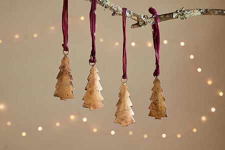 The Mishti Tree Baubles - Antique Brass - Small (Set of 4) by nkuku hang from a lichen-covered branch on burgundy ribbons, with a warm, softly blurred background and glowing lights for a festive, cozy feel.