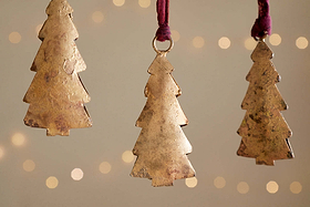 Four Mishti Tree Baubles in antique brass by nkuku hang on maroon ribbons against a soft, warm backdrop with blurred twinkling lights, adding a festive and cozy touch to your holiday decor.