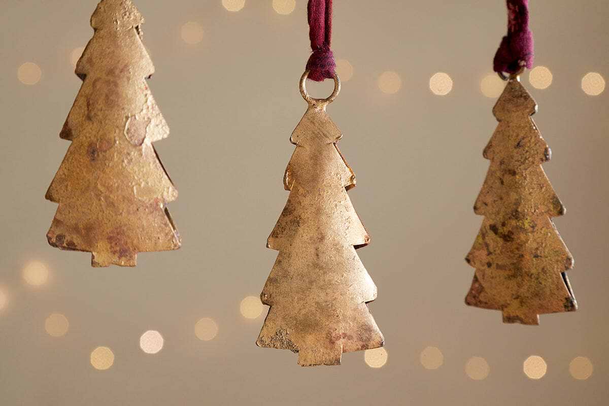 Four Mishti Tree Baubles in antique brass by nkuku hang on maroon ribbons against a soft, warm backdrop with blurred twinkling lights, adding a festive and cozy touch to your holiday decor.