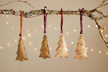 A lichen-covered branch displays four nkuku Mishti Tree Baubles in antique brass, each large bauble hung by maroon or orange ribbon. Warm white fairy lights blur softly behind, creating a charming festive scene.