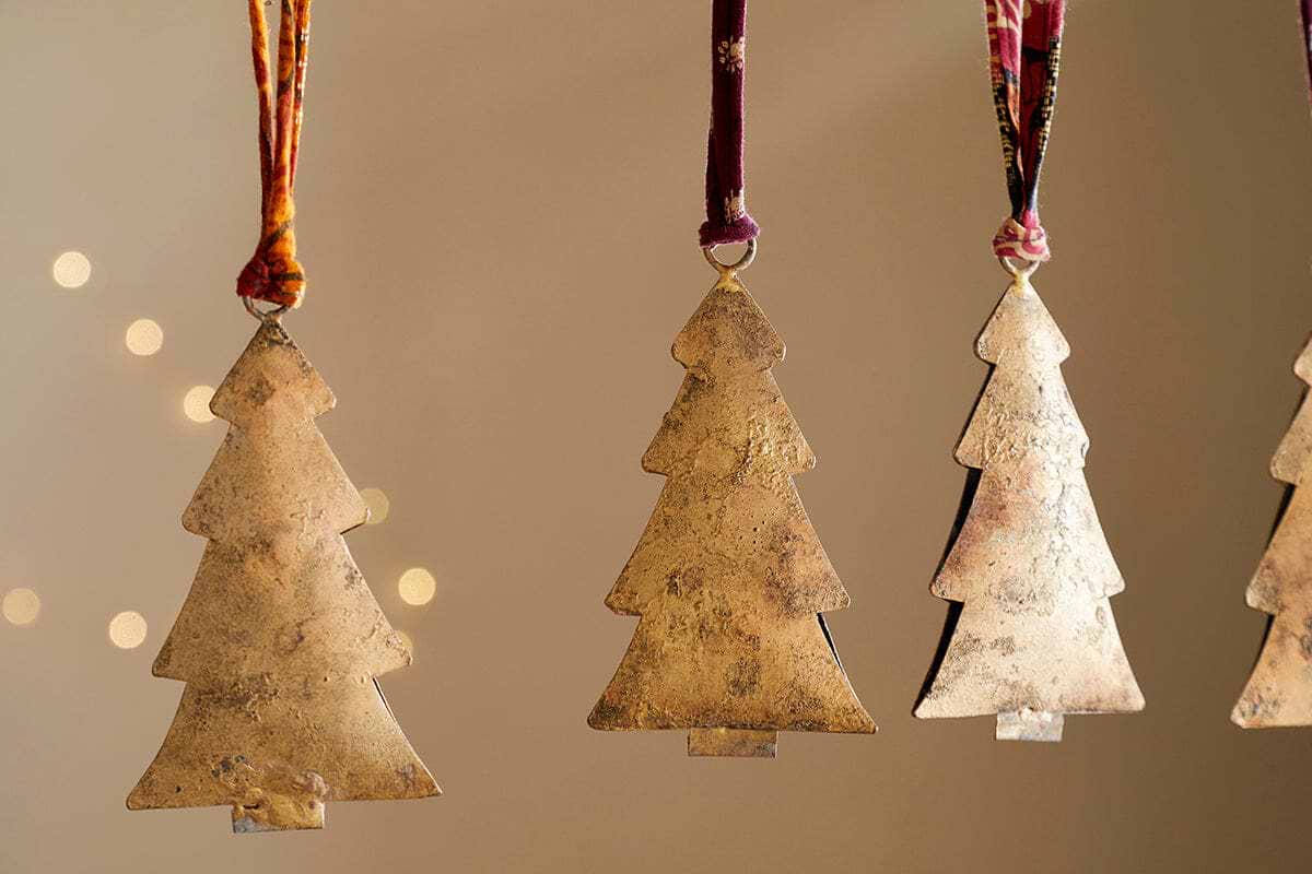 The nkuku Mishti Tree Baubles - Antique Brass - Large (Set of 4) are iron Christmas tree ornaments with a rustic, handcrafted look, hanging from ribbons against a neutral, softly lit background for a festive touch.