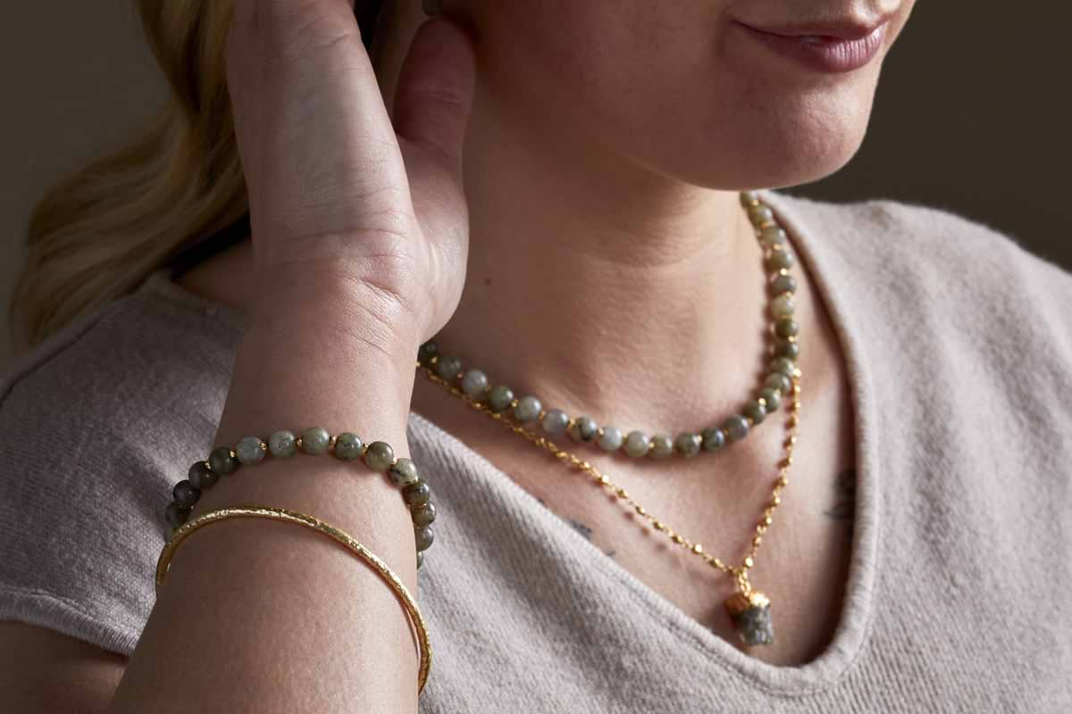 A woman wearing a light beige top and elegant jewellery, including nkuku’s Marai Bracelet – Labradorite, touches her ear with one hand.