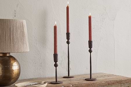 A wooden table features three lit red taper candles in nkuku Mawindi Candle Holders - Rust beside a round bronze lamp with a beige shade, set against a textured light wall for an earthy decor accent.