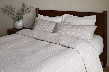 A neatly made bed featuring the nkuku Mora Hemp & Cotton Duvet Cover in Stone Grey, paired with two large pillows and a wooden headboard. A white vase with leafy branches sits on a bedside table against a neutral wall.