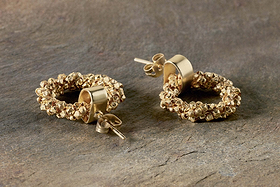 The nkuku Mula Earrings - Gold feature textured circular hoops made of faceted brass beads clustered on smooth cylindrical bases. Hypoallergenic and ideal for sensitive ears, they are shown on a stone-like surface.