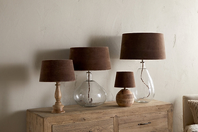 Four nkuku Mendai Velvet & Linen Lampshades in mocha are displayed on a rustic wooden cabinet. Three have transparent glass bases, and one has a textured ceramic base, set against a plain light-colored wall.