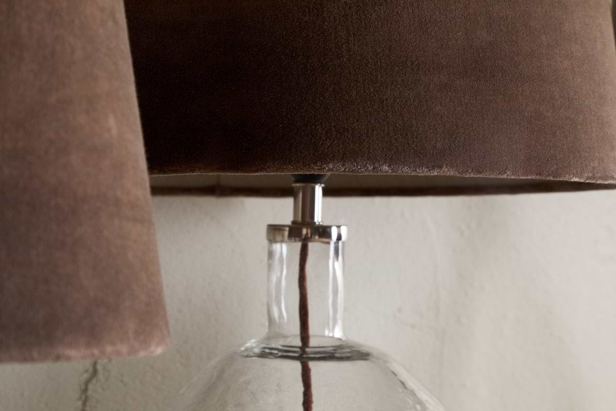 Close-up of the nkuku Mendai Velvet & Linen Lampshade in Mocha, paired with a clear glass lamp and visible cord, set against a textured white wall for a warm, inviting look.
