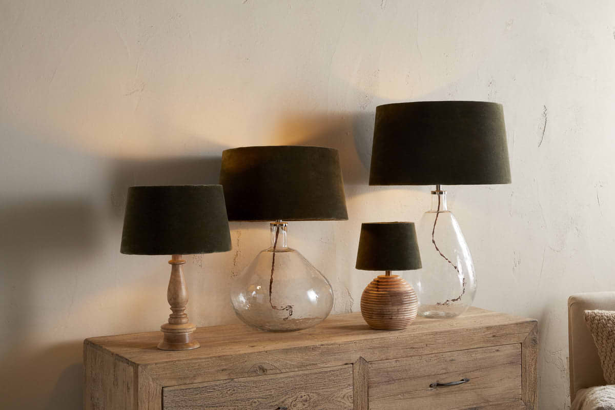 Four Mendai Velvet & Linen Lampshades in Moss Green by nkuku, with glass or ceramic bases, stand on a rustic wooden sideboard, casting soft, warm light and elegant shadows against a textured beige wall.