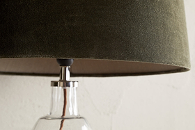 Close-up of the nkuku Mendai Velvet & Linen Lampshade in Moss Green, featuring a textured velvet finish. The glass base and cord are partially visible against a plain, light-toned wall that highlights the lamp's elegant details.