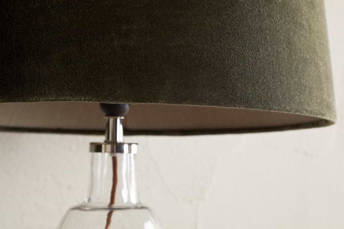 Close-up of the nkuku Mendai Velvet & Linen Lampshade in Moss Green, featuring a textured velvet finish. The glass base and cord are partially visible against a plain, light-toned wall that highlights the lamp's elegant details.