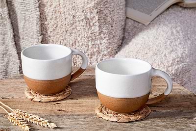 Two nkuku Mali White and Terracotta ceramic mugs rest on woven coasters atop a rustic wooden table. Paired with a soft throw and an open book, they create a cozy, inviting handmade pottery scene.