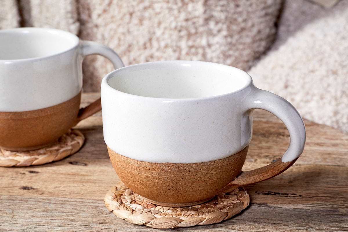 Two Mali White and Terracotta ceramic mugs by nkuku, featuring off-white glaze tops and terracotta bottoms, sit on woven coasters on a wooden table with a fuzzy blanket in the background.