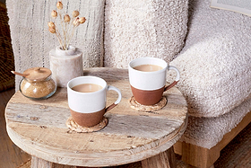 Two nkuku Mali White & Terracotta mugs with milky coffee rest on woven coasters on a rustic table, beside a brown sugar jar with a wooden lid and a ceramic vase of dried flowers. A textured beige armchair is partly visible.