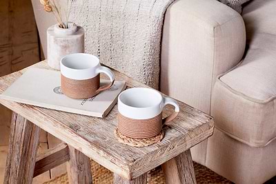 The nkuku Mali Ribbed Coffee Mug - White (Set of 2) adds artisan charm to your cozy corner when placed on a rustic stool beside a beige armchair with a textured throw blanket.
