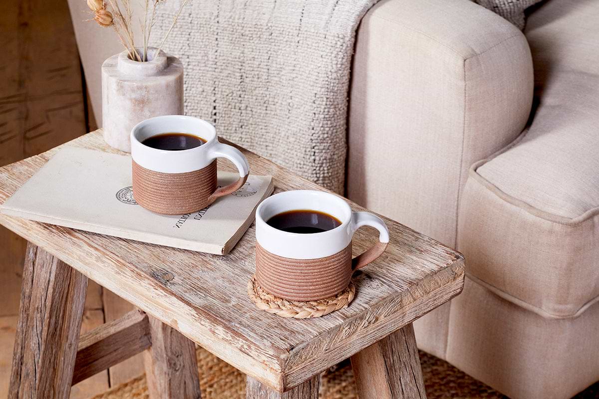 Two nkuku Mali Ribbed Coffee Mugs in white (set of 2), one filled with black coffee, sit on a rustic wooden table next to a beige envelope, a vase of dried flowers, and a textured light beige sofa.