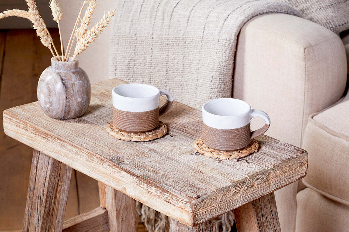 A rustic wooden table displays two nkuku Mali Ribbed Espresso Mugs - White on woven coasters. A textured vase with dried wheat, beige armchairs, and a cozy knit blanket add warmth and inviting style to the scene.
