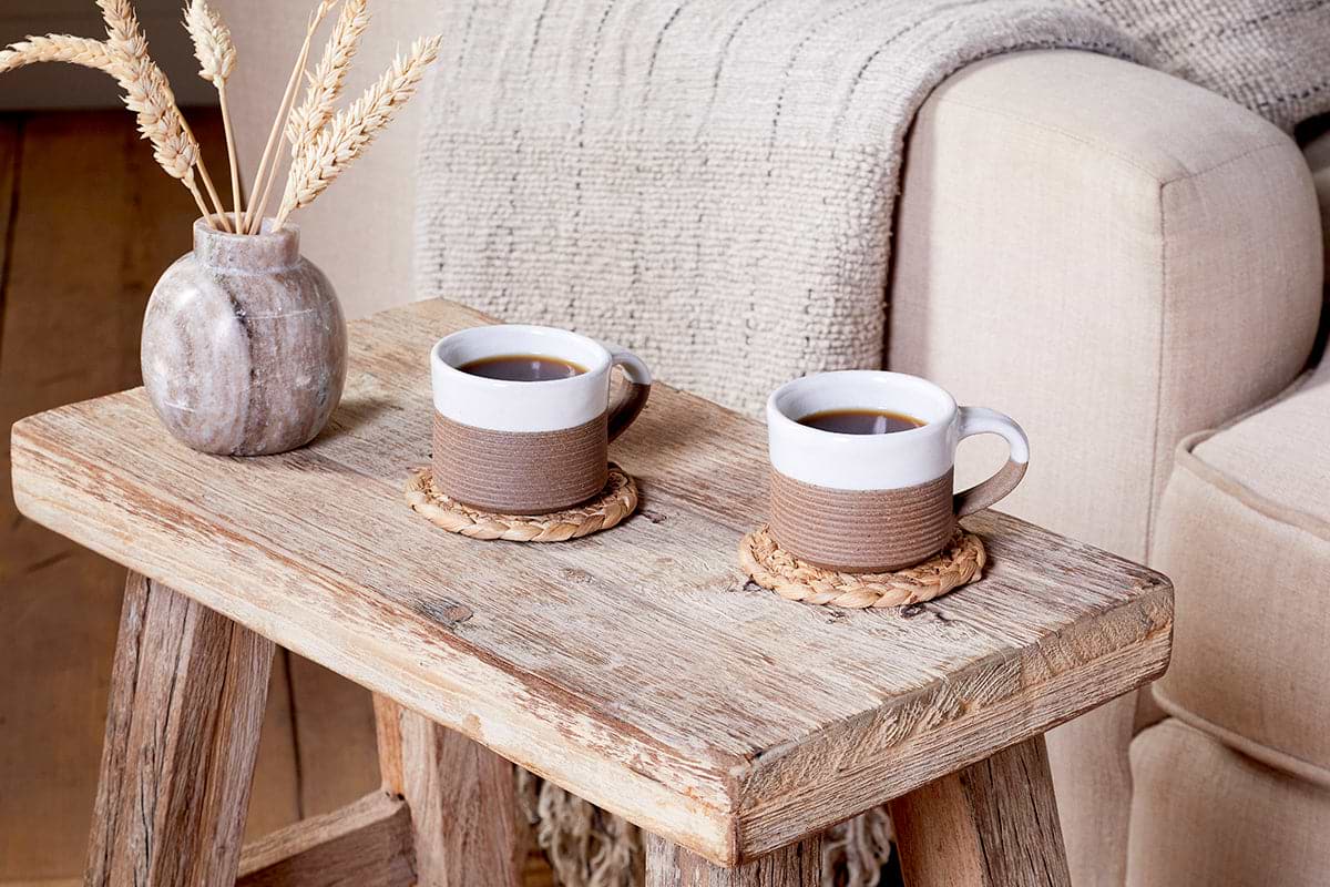 Two nkuku Mali Ribbed Espresso Mugs in white are filled with coffee and placed on woven coasters on a rustic wood table. A textured vase with dried grasses and a beige knit blanket over a cream sofa create a cozy, welcoming space.