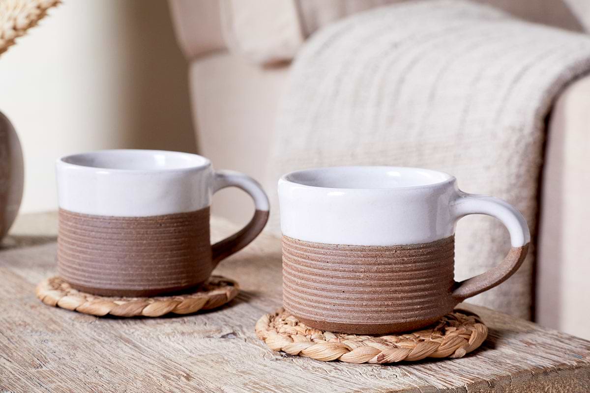 The nkuku Mali Ribbed Espresso Mug - White (Set of 2) rests on woven coasters atop a wooden table, with a beige knitted blanket draped over a sofa in the background for a cozy, inviting feel.