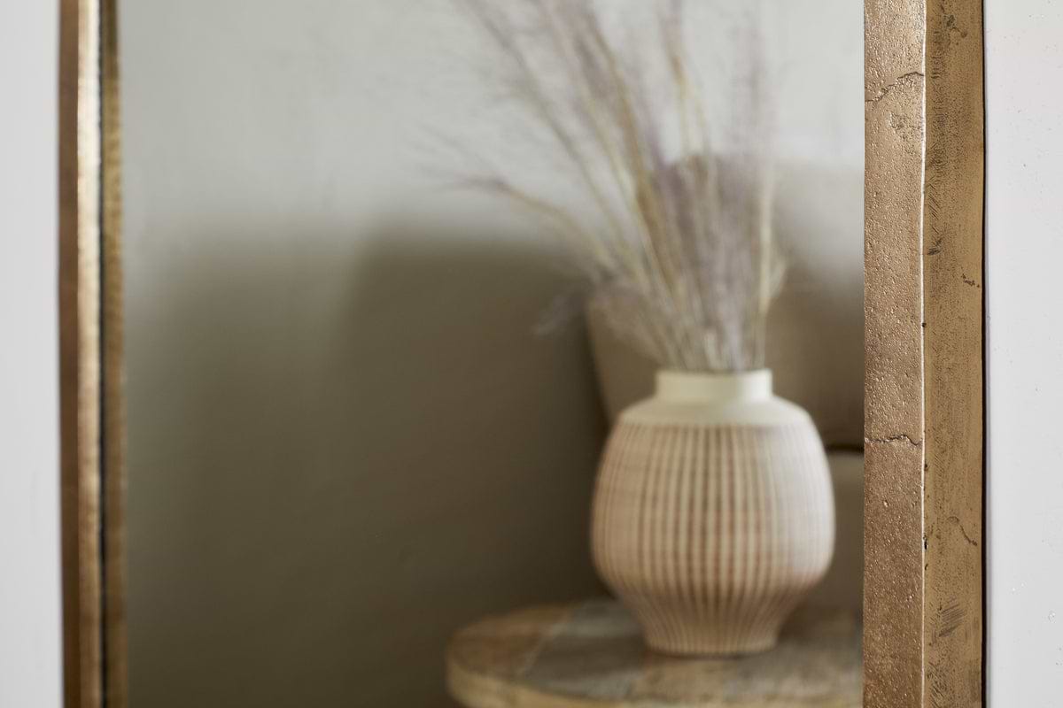 The Murwara Full Length Mirror in antique brass by nkuku reflects a ribbed ceramic vase with dried grasses, resting on a round light tabletop in a softly lit, neutral room for a minimalist, elegant look.